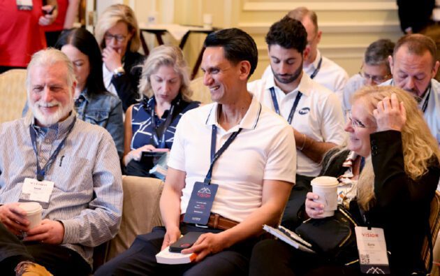 A group of people with conference badges gather in a meeting room, discussing workforce management over coffee, while some smile and others review their phones or notes.