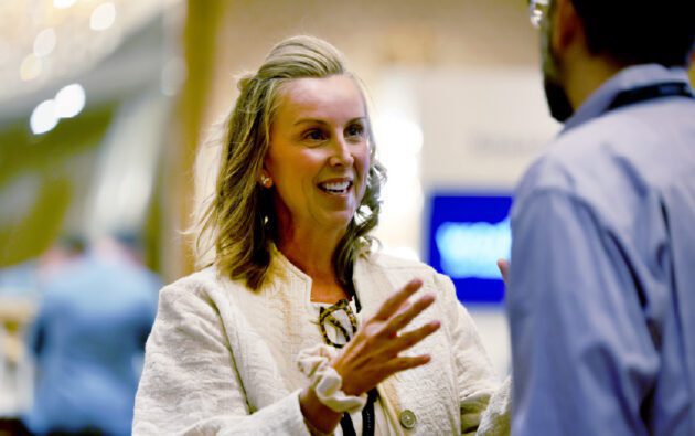 A woman in a light-colored jacket gestures while discussing workforce management with a man in a blue shirt at an indoor event.