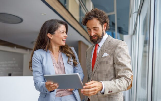 Two business professionals, a woman and a man, stand indoors by a window, smiling and looking at a tablet device together.