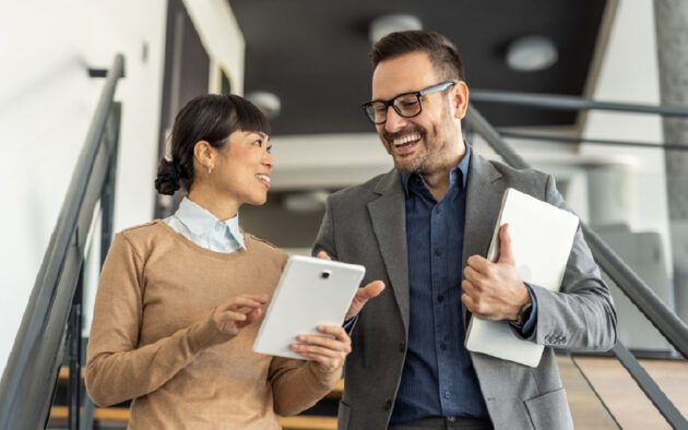 Two professionals stand on stairs, smiling and conversing. One holds a tablet and the other holds a laptop.
