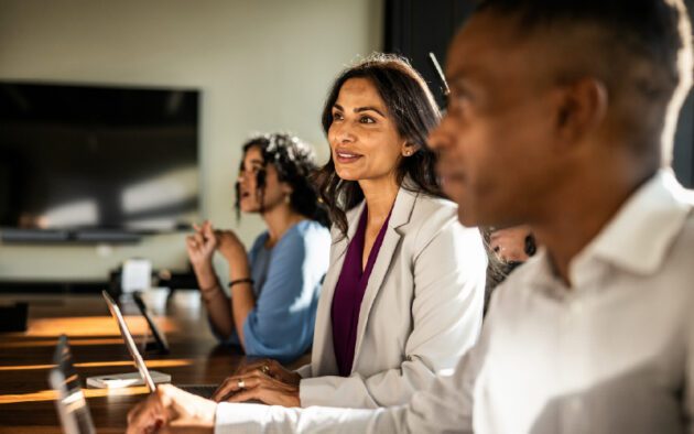 Three professionals sit at a conference table, attentively listening during a meeting, with laptops and notepads in front of them.
