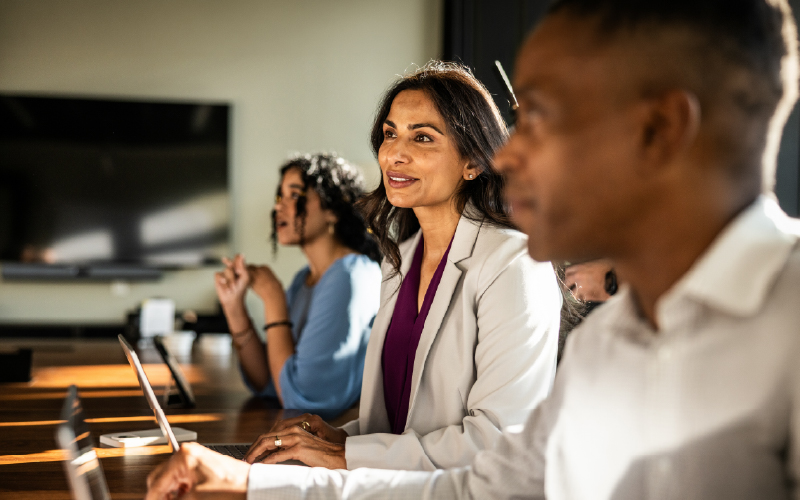 Three professionals sit at a conference table, attentively listening during a meeting, with laptops and notepads in front of them.