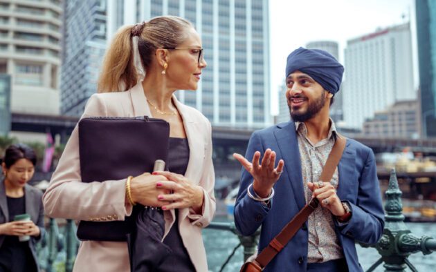 A woman and a man in business attire walk and converse on a city street, with office buildings in the background.