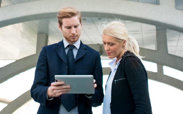 Two business professionals standing outdoors, looking at a tablet together under a modern architectural structure.