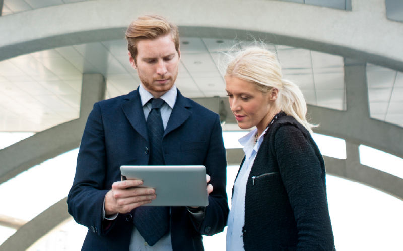 Two business professionals standing outdoors, looking at a tablet together under a modern architectural structure.