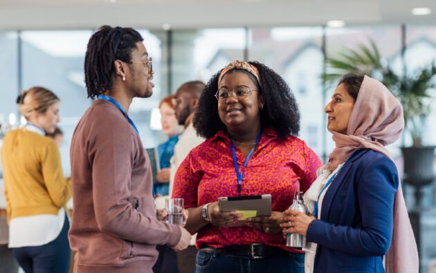 Three people wearing conference badges stand together and talk, while holding drinks and a tablet, with other attendees interacting in the background.