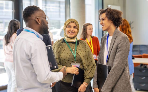 A group of professionals stand and talk in an office setting, holding laptops and wearing conference badges.