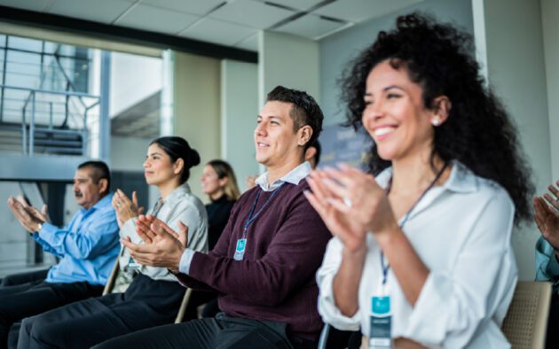 A group of people sitting in chairs and clapping, likely attending a seminar or conference in a modern indoor setting.