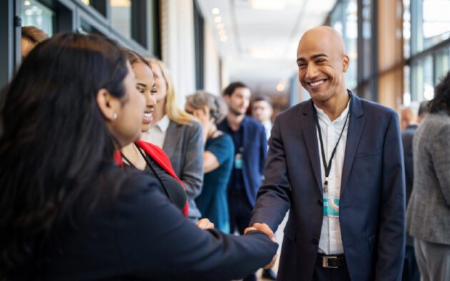 A man in a suit smiles and shakes hands with a woman at a professional networking event with other people in the background.
