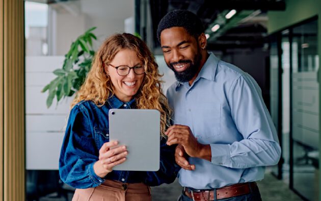Two colleagues stand indoors, smiling and looking at a tablet device together.