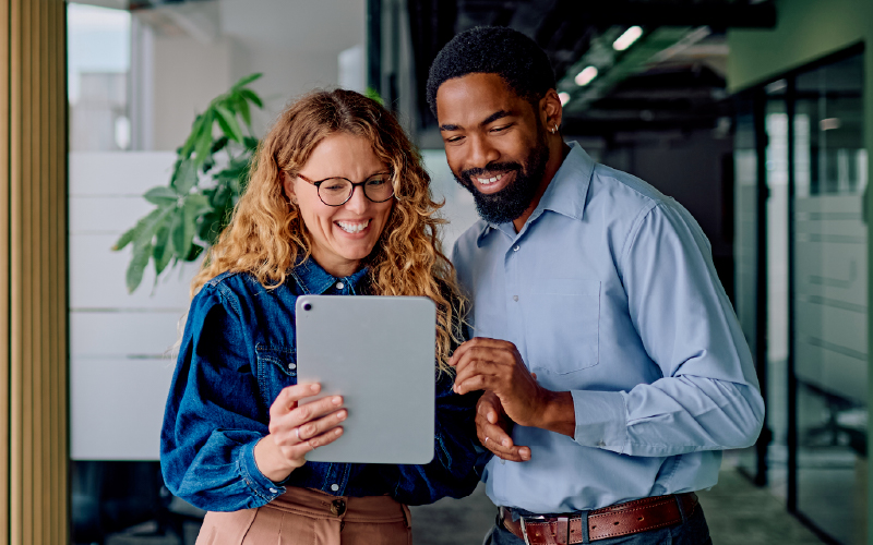 Two colleagues stand indoors, smiling and looking at a tablet device together.
