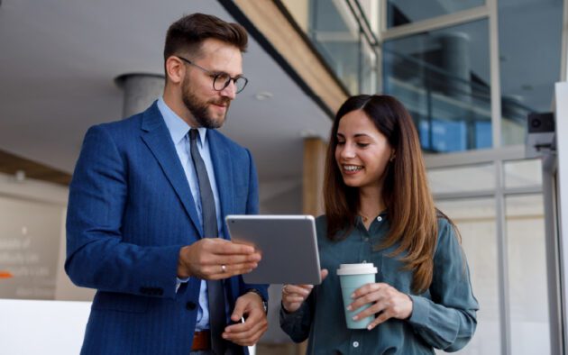 A man in a suit shows a tablet to a woman holding a coffee cup; both are standing indoors and appear to be having a discussion.
