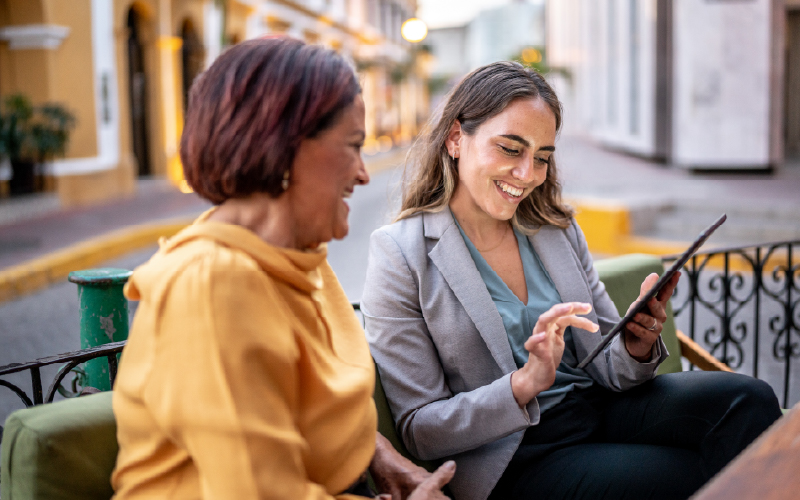 Two women sit outdoors on a bench, smiling as one shows the other something on a tablet.