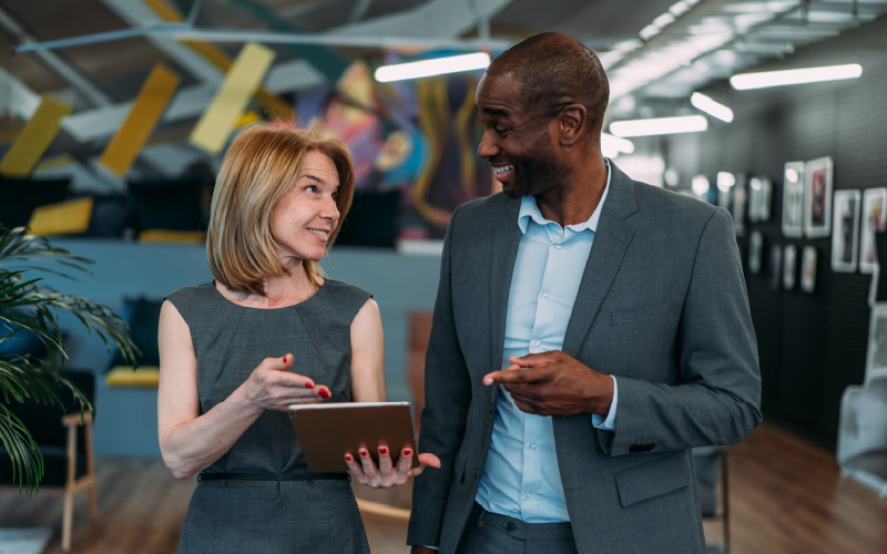 Two business professionals in gray suits stand indoors, smiling and conversing while one holds a tablet.