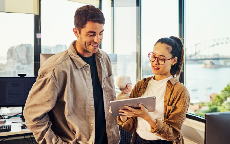 Two people stand in a modern office by a window, looking at a tablet together. A cityscape and a bridge are visible outside.