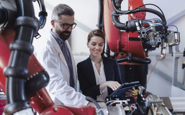 Two professionals examine machinery in a modern industrial setting; one holds a tablet, while robotic arms and equipment are visible around them.