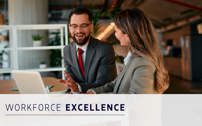 Two business professionals in suits sit at a table with a laptop, smiling and talking. Text overlay reads "Workforce Excellence." Modern office setting with plants in the background.