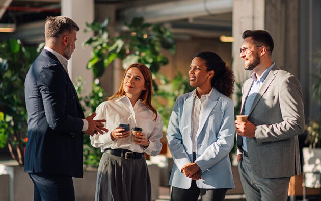 Four people in business attire stand together indoors, holding cups and talking in a modern office space with large plants in the background.