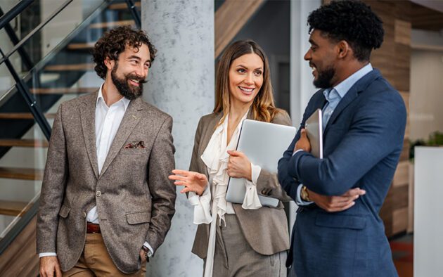 Three professionals in business attire stand indoors, holding laptops and papers, conversing and smiling near a staircase in a modern office setting.