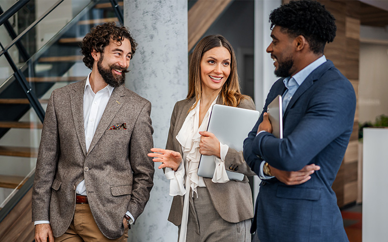 Three professionals in business attire stand indoors, holding laptops and papers, conversing and smiling near a staircase in a modern office setting.
