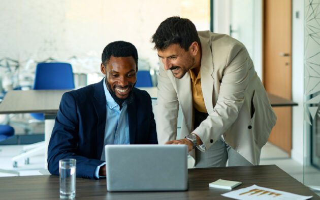 Two men in business attire look at a laptop screen and smile in a modern office setting, with papers and a glass of water on the table.