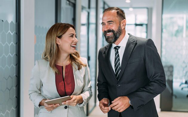 Two business professionals, a woman holding a tablet and a man in a suit, are walking and smiling in a modern office hallway.