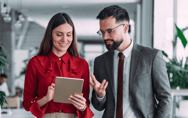 Two professionals, one holding a tablet, discuss workforce management governance while standing in a modern office with large windows and plants.