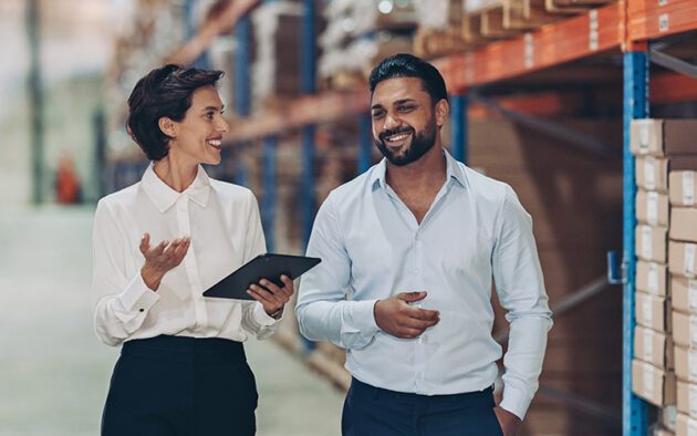 Two people in business attire walk and talk in a warehouse aisle, with shelves of boxes in the background. One holds a digital tablet.