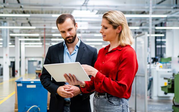 Two people stand in a factory or industrial setting, looking at a tablet together. The woman is holding the tablet and pointing at the screen while the man watches attentively.