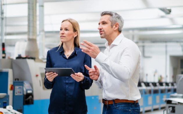 A man and woman standing in a manufacturing facility, with the man gesturing as he speaks to the woman holding a tablet.