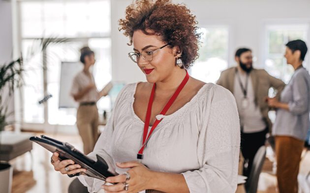 A woman in a white blouse and red lanyard holds a tablet in an office setting with coworkers in the background.