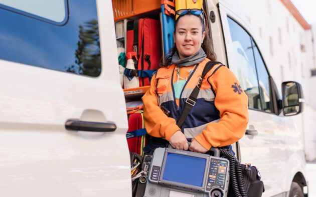 A female paramedic in an orange and blue uniform, dressed in accordance with workforce regulations, stands by an open ambulance holding a portable medical monitor.