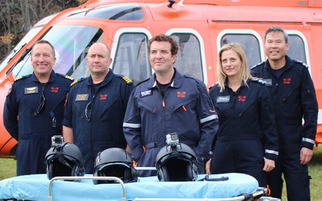 Five Ornge air ambulance crew members in dark blue uniforms stand in front of an orange helicopter behind a medical gurney with three flight helmets.