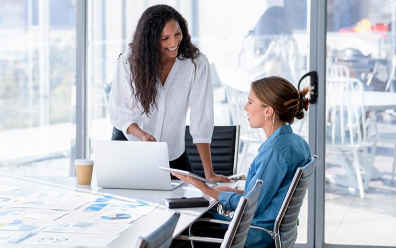 Two women engaged in a discussion at a desk with a laptop and documents in a bright office setting.