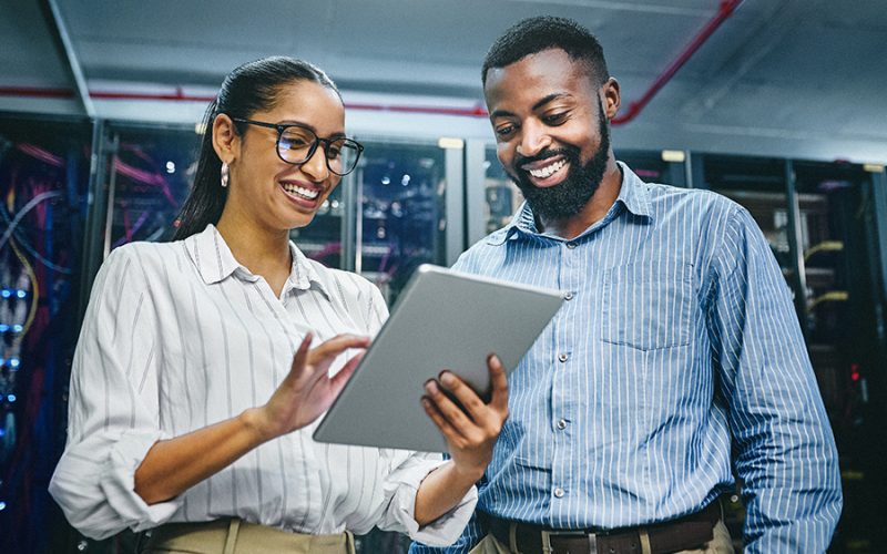 Two people smiling while looking at a tablet in a server room.