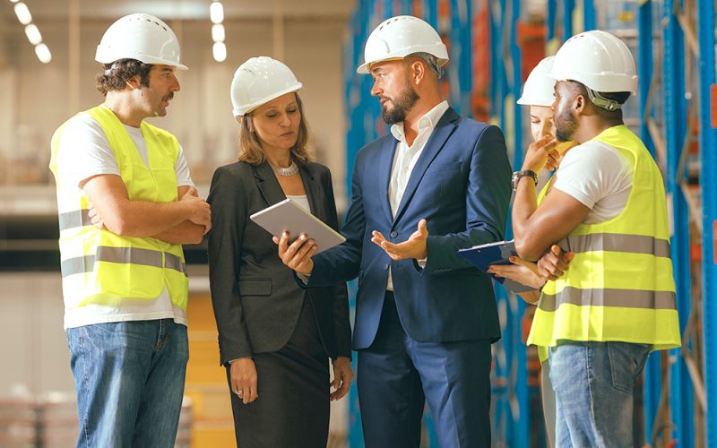 A group of five people, including three workers in safety vests and helmets, and two individuals in formal attire, stand discussing something in a warehouse. One person is holding a tablet.