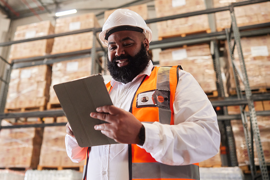 A warehouse worker in a safety vest and hard hat uses a tablet while standing in front of shelves stacked with boxes.